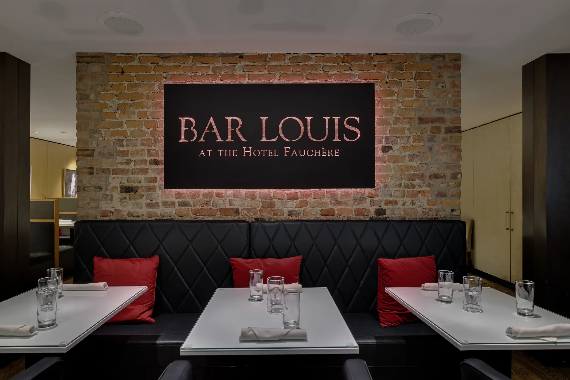 Elegant seating at Bar Louis featuring red pillows and white tables against a rustic brick wall in Milford, PA.