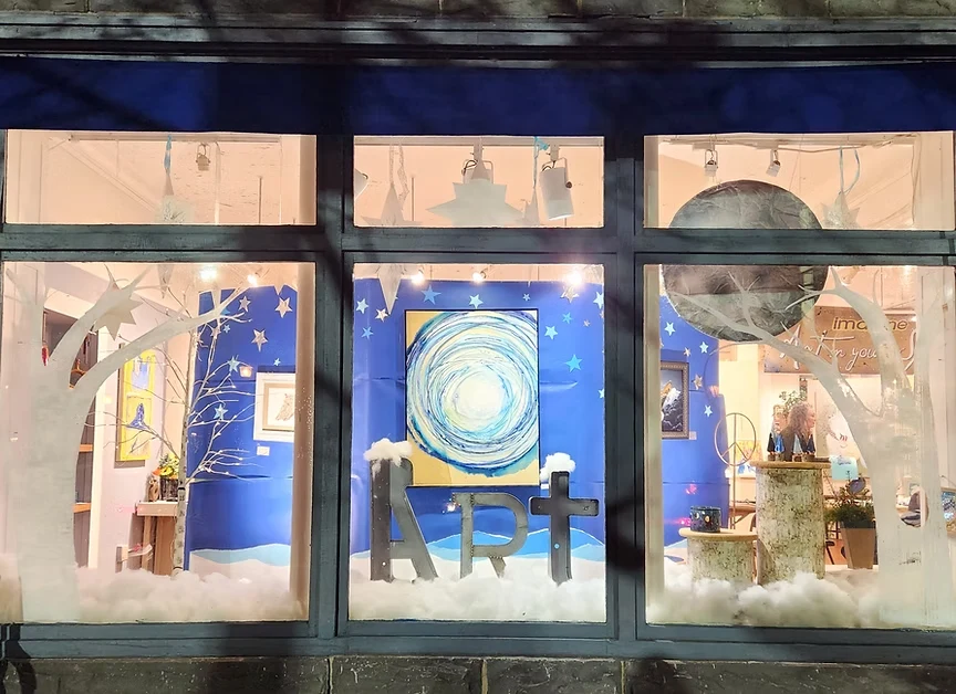 An illuminated storefront window in Milford PA shows local art happenings with a celestial night sky theme.