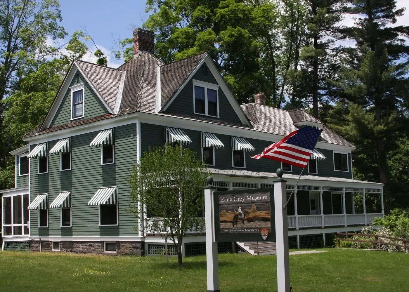 The Zane Grey Museum features green siding and striped window awnings beneath an American flag in Milford, PA.