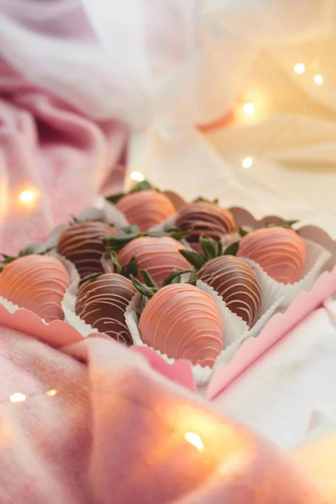 A box of drizzled chocolate-covered strawberries sits on a soft pink fabric surrounded by warm fairy lights.