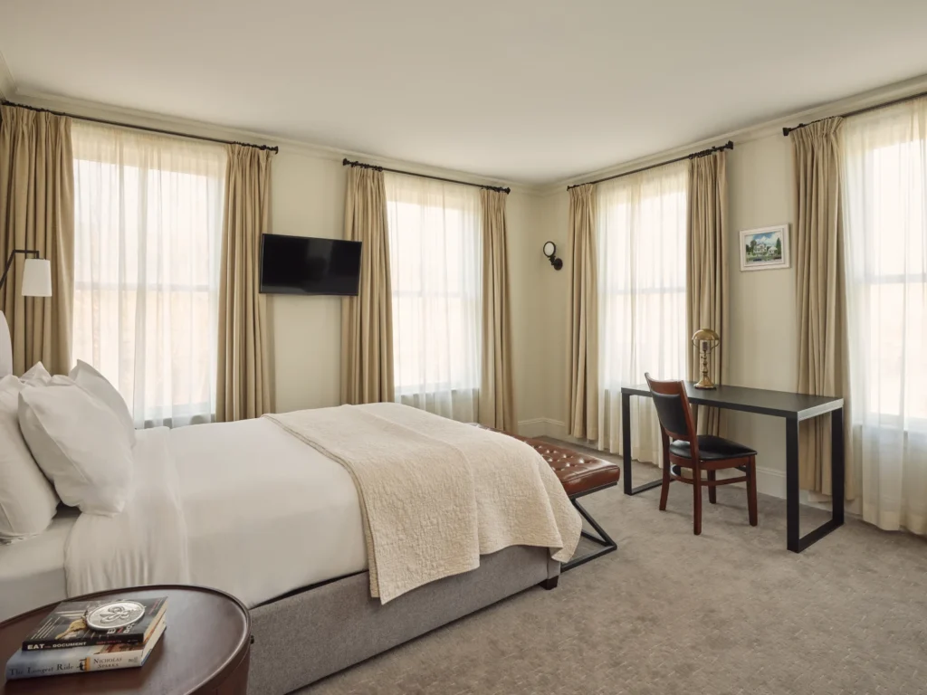 A sunlit guest room at Hotel Fauchère features plush white bedding, a study desk, and large curtained windows.