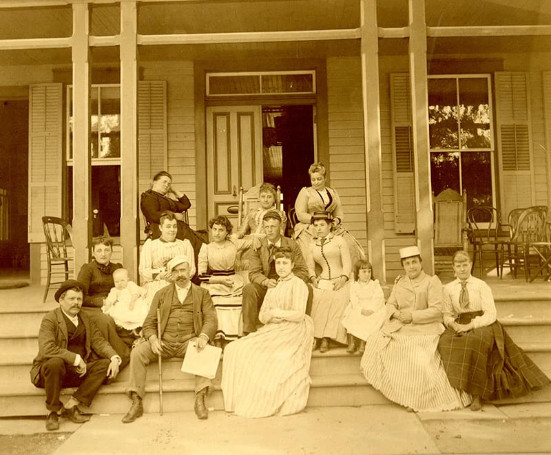 A vintage sepia photo of a group in Victorian attire sitting on the porch of the historic Hotel Fauchère.